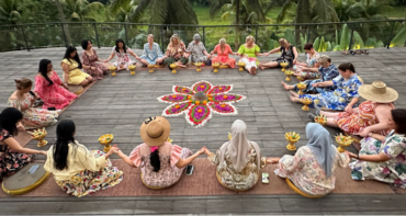 A diverse group of women holding hands in a large, supportive circle around a vibrant flower mandala ceremony during the Curves Bali Retreat at Soulshine Ubud.