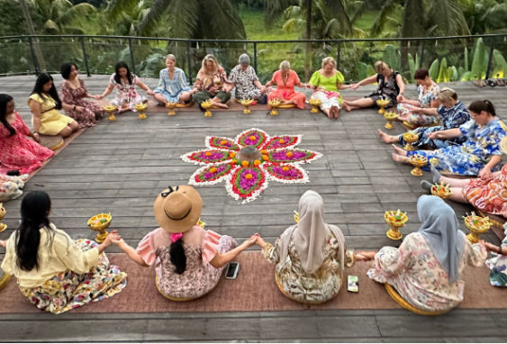 A diverse group of women holding hands in a large, supportive circle around a vibrant flower mandala ceremony during the Curves Bali Retreat at Soulshine Ubud.