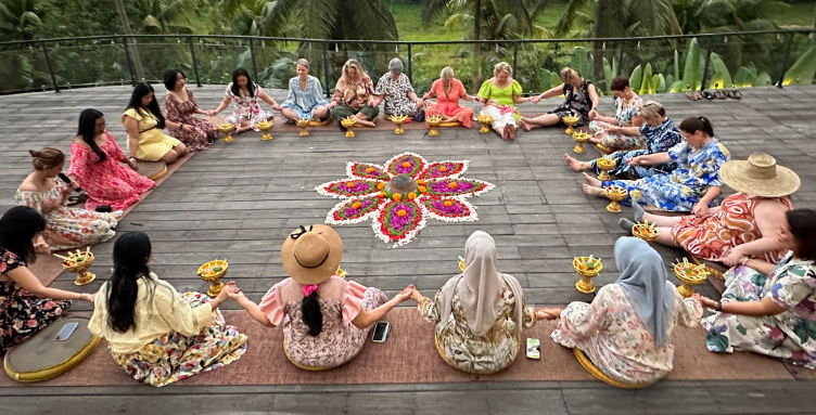 A diverse group of women holding hands in a large, supportive circle around a vibrant flower mandala ceremony during the Curves Bali Retreat at Soulshine Ubud.