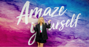 Curves CEO Kim McQueen smiling playfully in front of a purple and blue watercolor wall that reads 