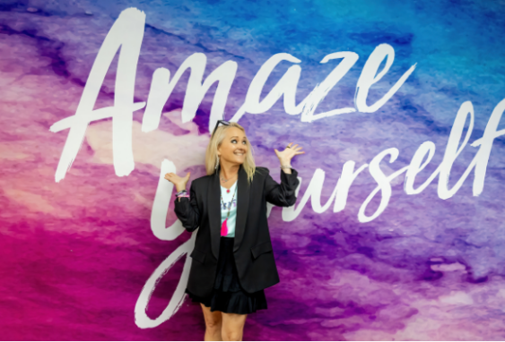 Curves CEO Kim McQueen smiling playfully in front of a purple and blue watercolor wall that reads 