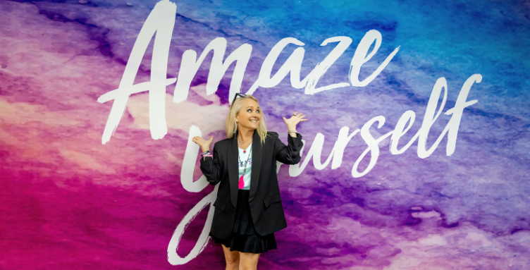 Curves CEO Kim McQueen smiling playfully in front of a purple and blue watercolor wall that reads 