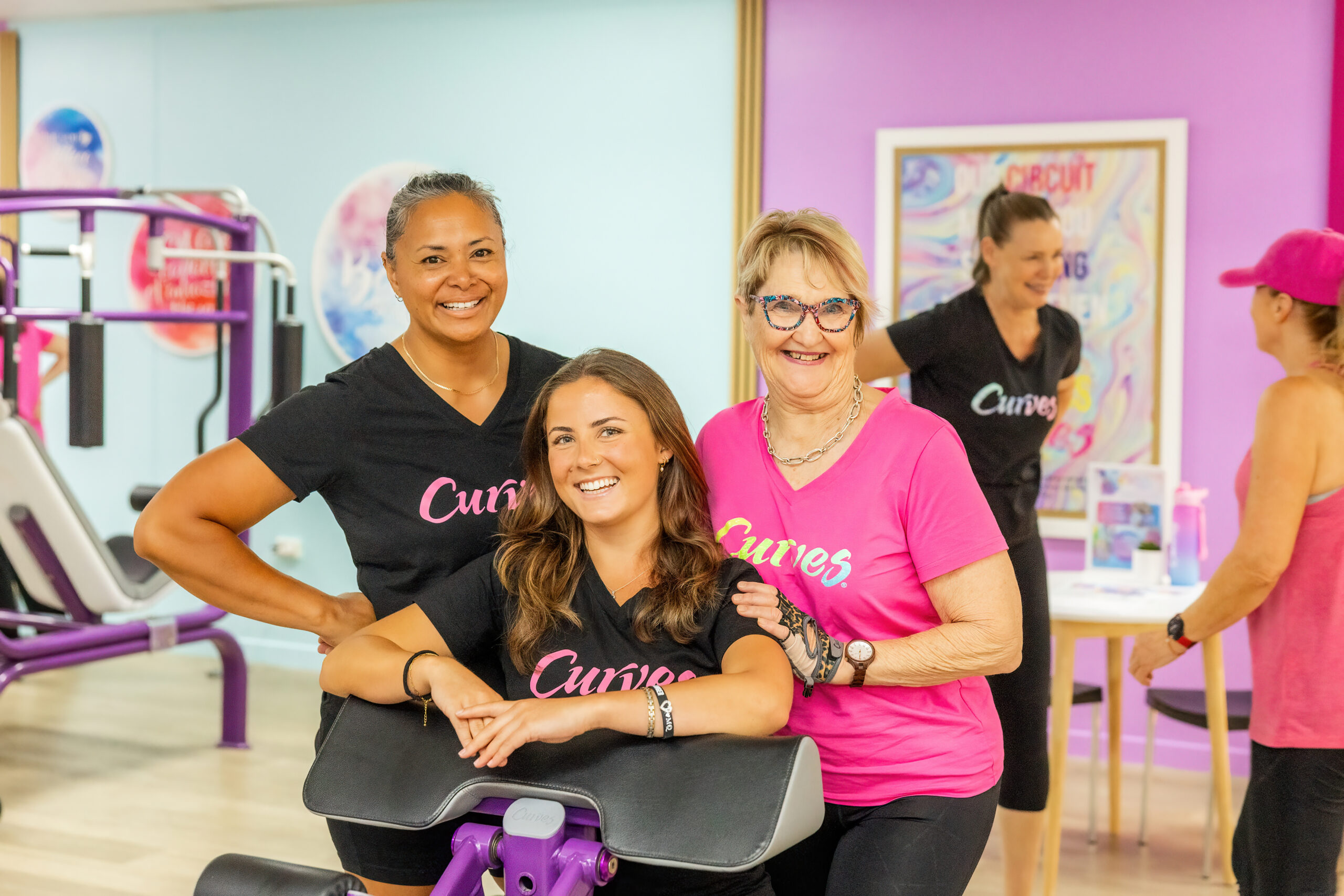 A group of cheerful women smiling inside a bright Curves fitness club. In the foreground, three women pose closely together by a purple workout machine; two wear textured black Curves v-neck t-shirts and one wears a bright pink Curves t-shirt. In the soft-focus background, another coach in a black Curves shirt stands near a colorful "Circuit" poster, while a woman in a pink cap and tank top stands by a small table.