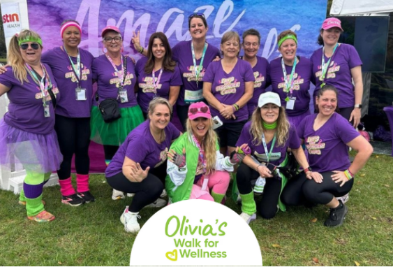 • A large group of women wearing matching purple Curves fitness shirts stand and crouch together outside near the official event tent at Olivia's Walk for Wellness.