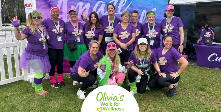 • A large group of women wearing matching purple Curves fitness shirts stand and crouch together outside near the official event tent at Olivia's Walk for Wellness.