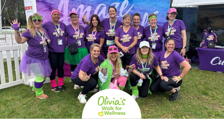 • A large group of women wearing matching purple Curves fitness shirts stand and crouch together outside near the official event tent at Olivia's Walk for Wellness.
