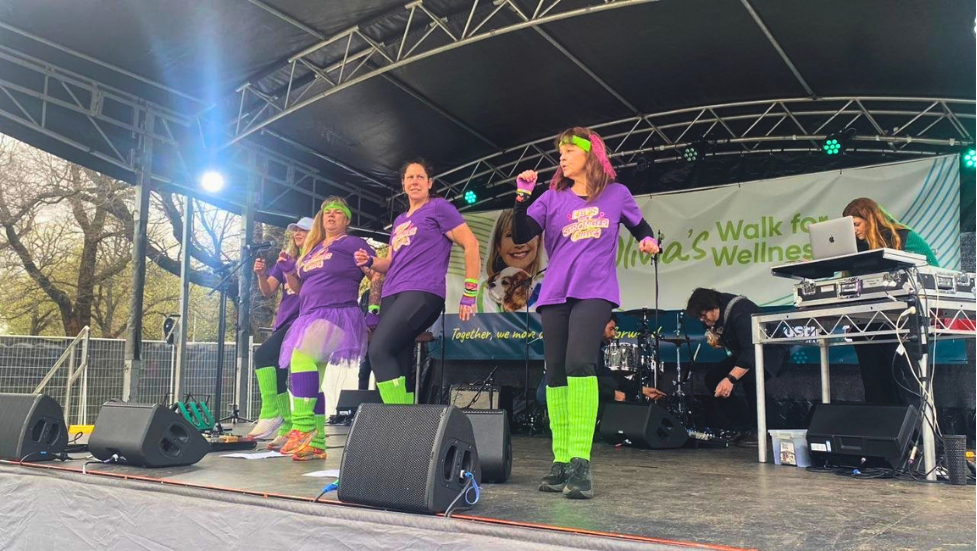 A group of Curves women in purple shirts and bright neon green leg warmers lead an energetic exercise routine on an outdoor stage at the ONJ Walk for Wellness 2026
