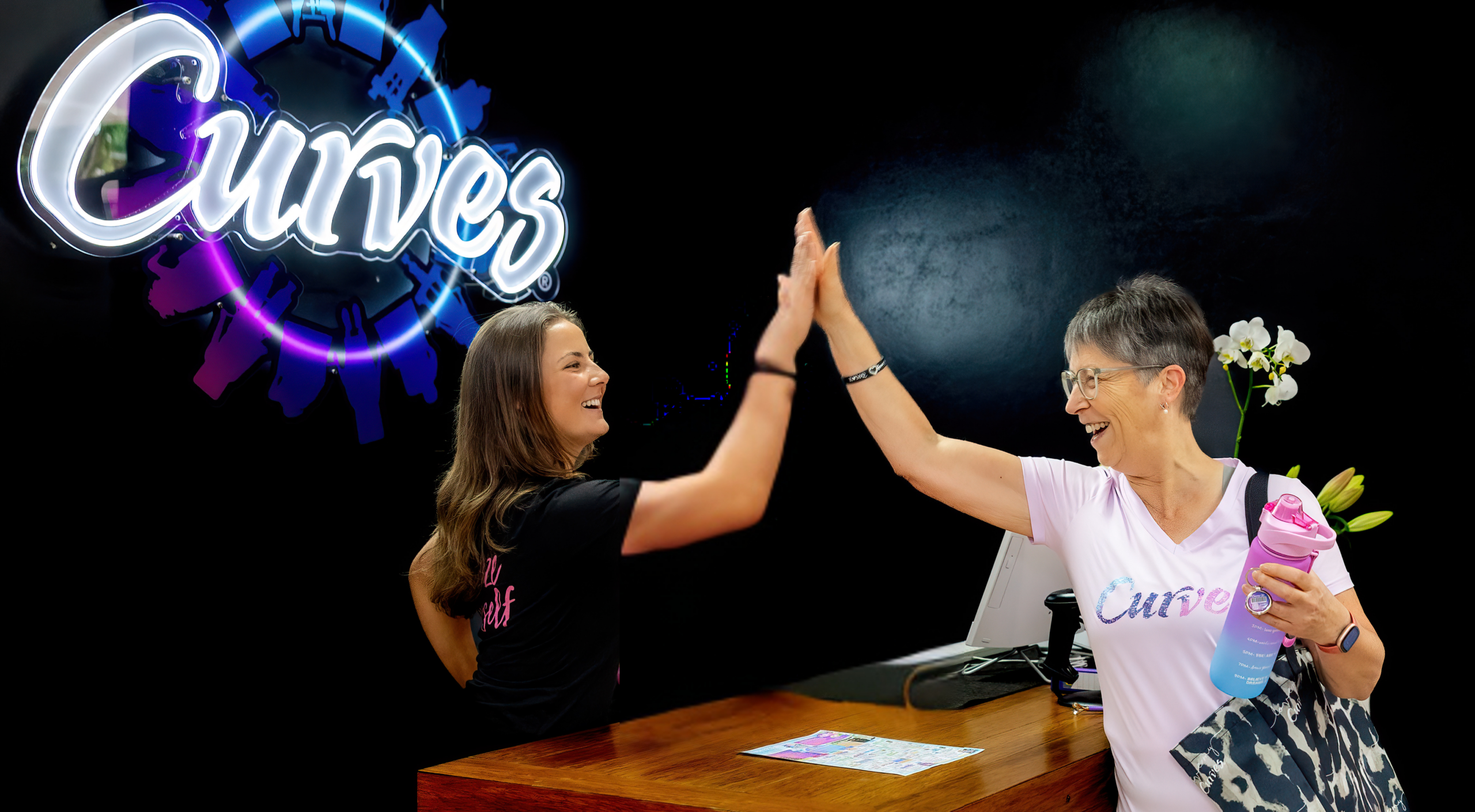 A smiling Curves coach behind a wooden reception desk gives a high-five to a cheerful mature member. The member is wearing a white Curves t-shirt, carrying a patterned bag, and holding a pink and blue gradient water bottle. A glowing purple and blue neon Curves logo shines on the black wall behind them.